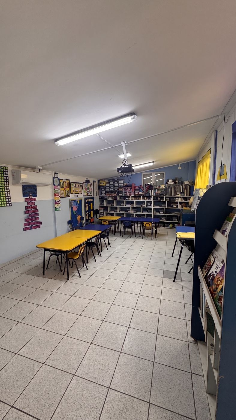 Sala de prekínder, con mesas amarillas, biblioteca de aula y murales